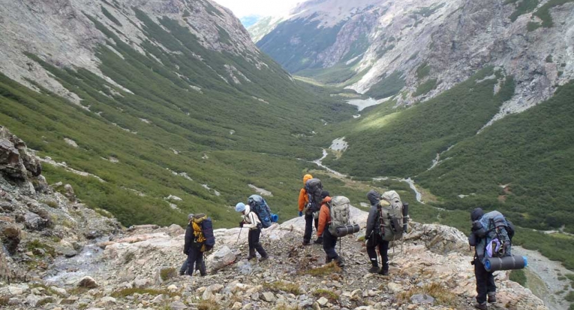 A group of people wearing backpacks hike over a rocky cliff, overlooking a vast green valley surrounded by mountains. There is a narrow river winding through the valley. 